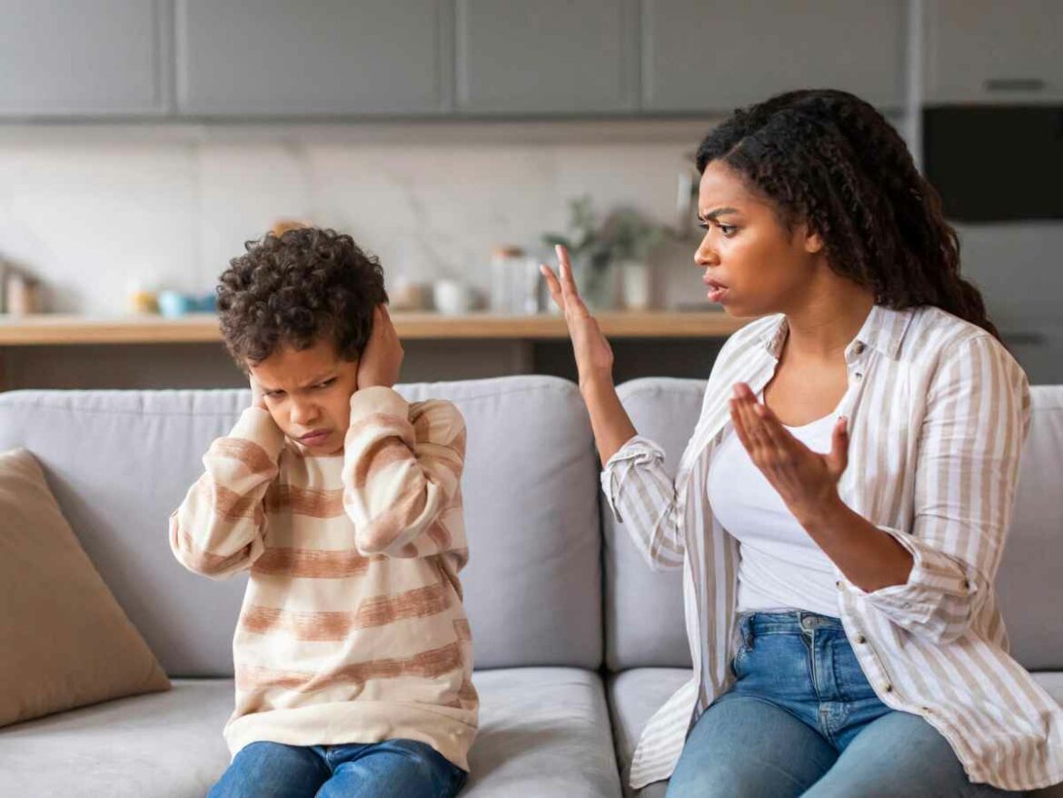 Mulher repreende uma criança em casa; o menino, com expressão de desconforto, cobre os ouvidos com as mãos.