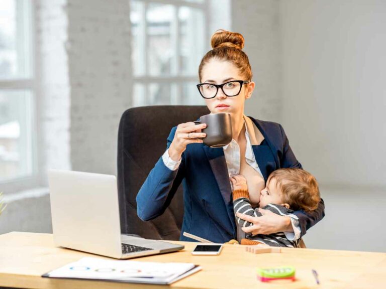 Mãe amamenta o bebê no colo enquanto trabalha em casa, vestida com roupa social e segurando uma caneca, representando a conciliação entre maternidade e carreira.