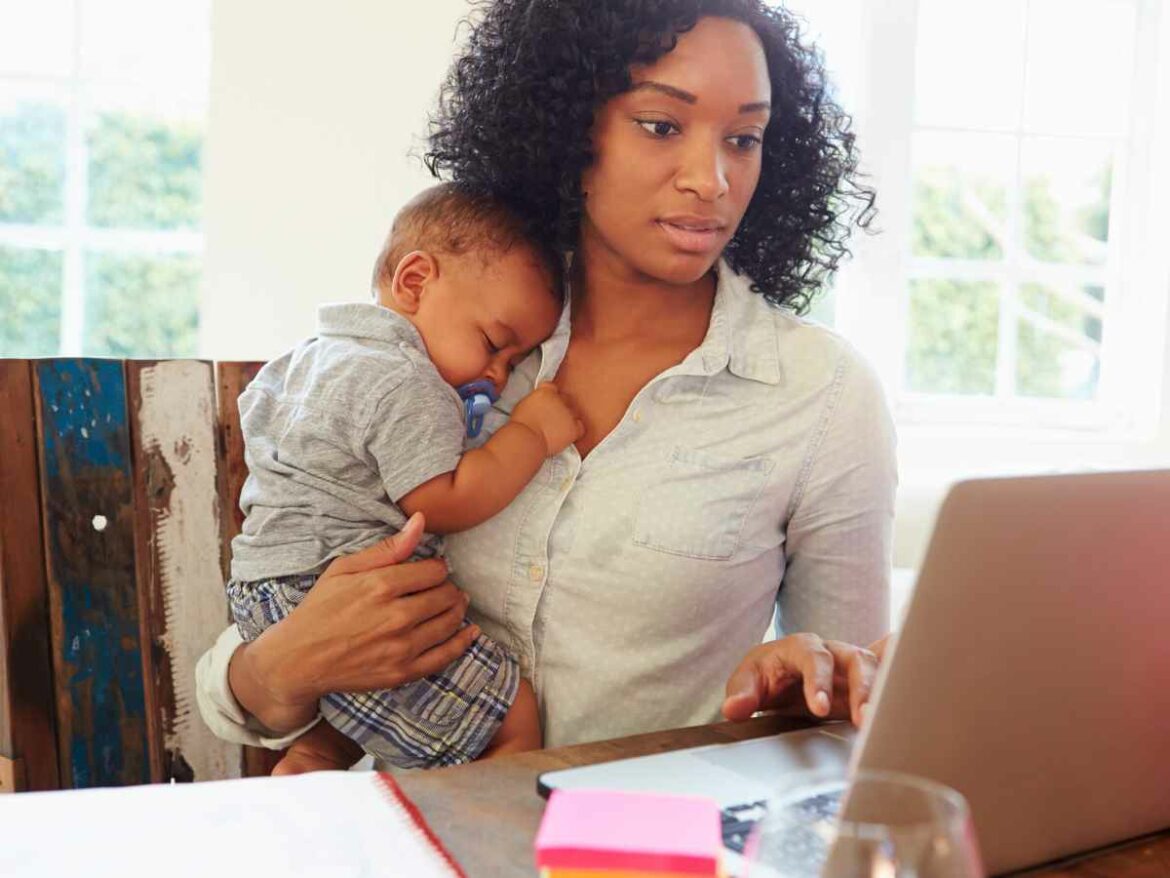 Mãe com expressão concentrada segura o bebê adormecido no colo enquanto trabalha no computador, representando o momento de organização logística para o retorno ao trabalho após a licença-maternidade.