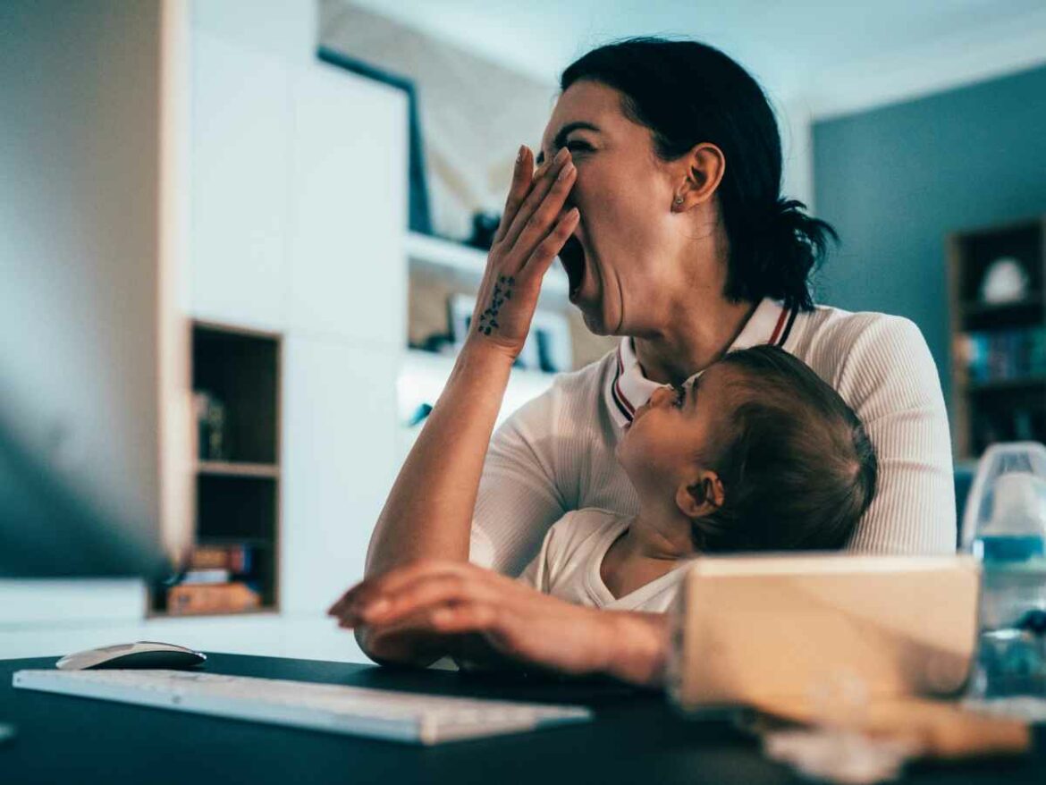 Mãe visivelmente cansada boceja enquanto segura o bebê no colo e trabalha no computador, simbolizando os desafios da exaustão no retorno à rotina após a licença-maternidade.
