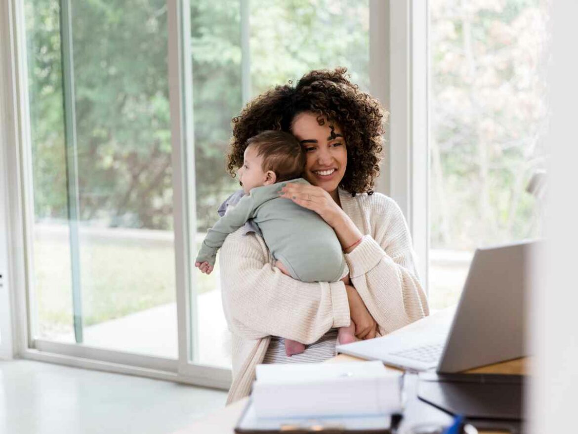 Mãe sorridente segura o bebê no colo enquanto trabalha em home office, representando a adaptação da rotina com afeto e equilíbrio após a volta ao trabalho.