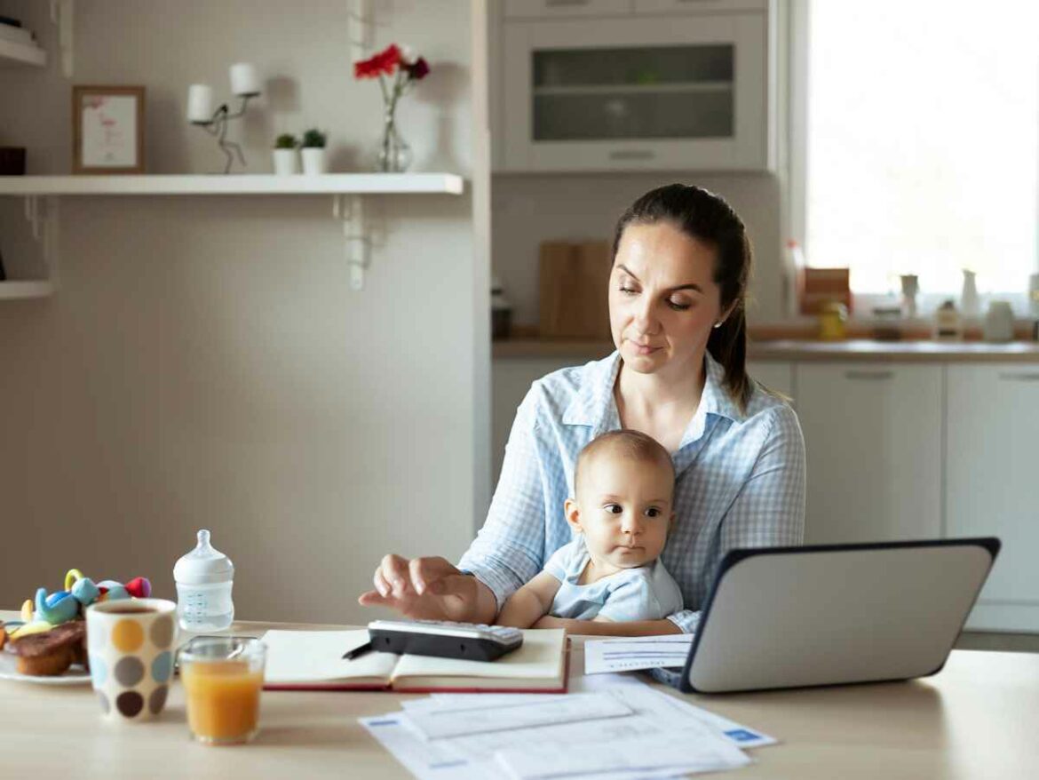 Mãe com bebê no colo organiza contas e agenda em frente ao notebook na cozinha, simbolizando o planejamento da rotina após o término da licença-maternidade.