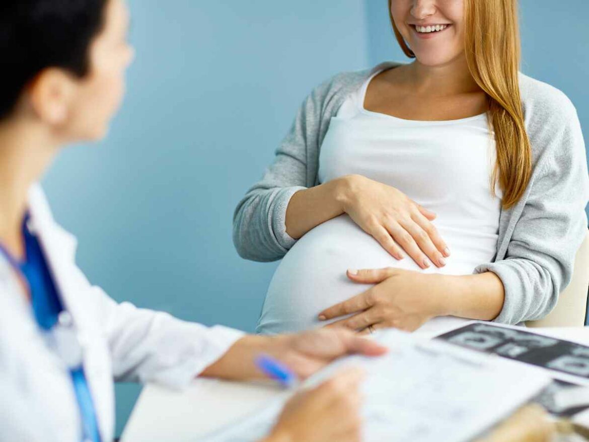 Gestante sorridente durante consulta de pré-natal com profissional da saúde, segurando a barriga com uma das mãos.