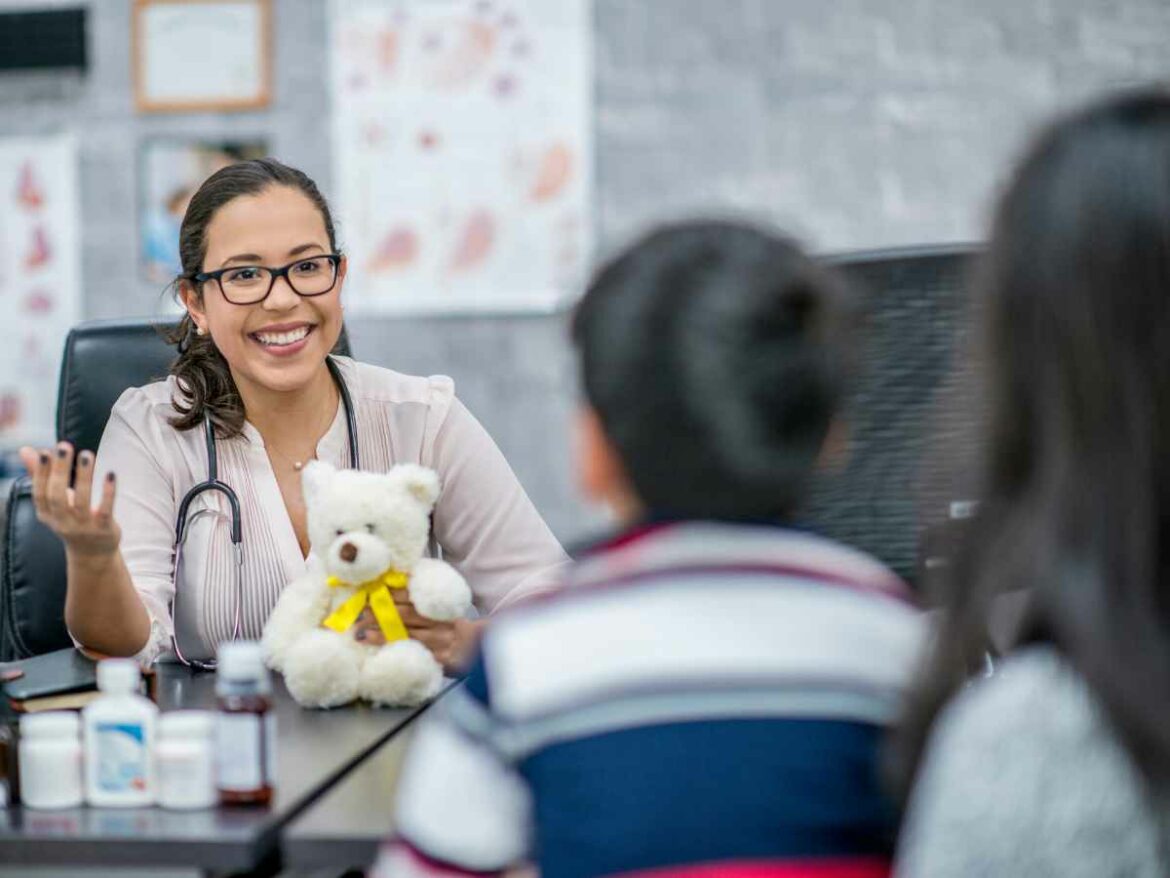 Pediatra sorridente conversa com pais e filho durante consulta, segurando um ursinho de pelúcia. A cena transmite acolhimento e confiança.
