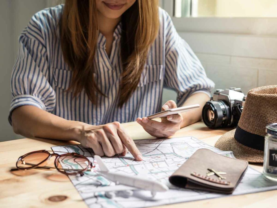 Mulher apontando um destino em um mapa enquanto segura o celular, com itens de viagem ao redor, representando o planejamento cuidadoso de uma viagem.