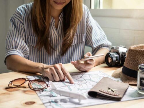 Mulher apontando um destino em um mapa enquanto segura o celular, com itens de viagem ao redor, representando o planejamento cuidadoso de uma viagem.