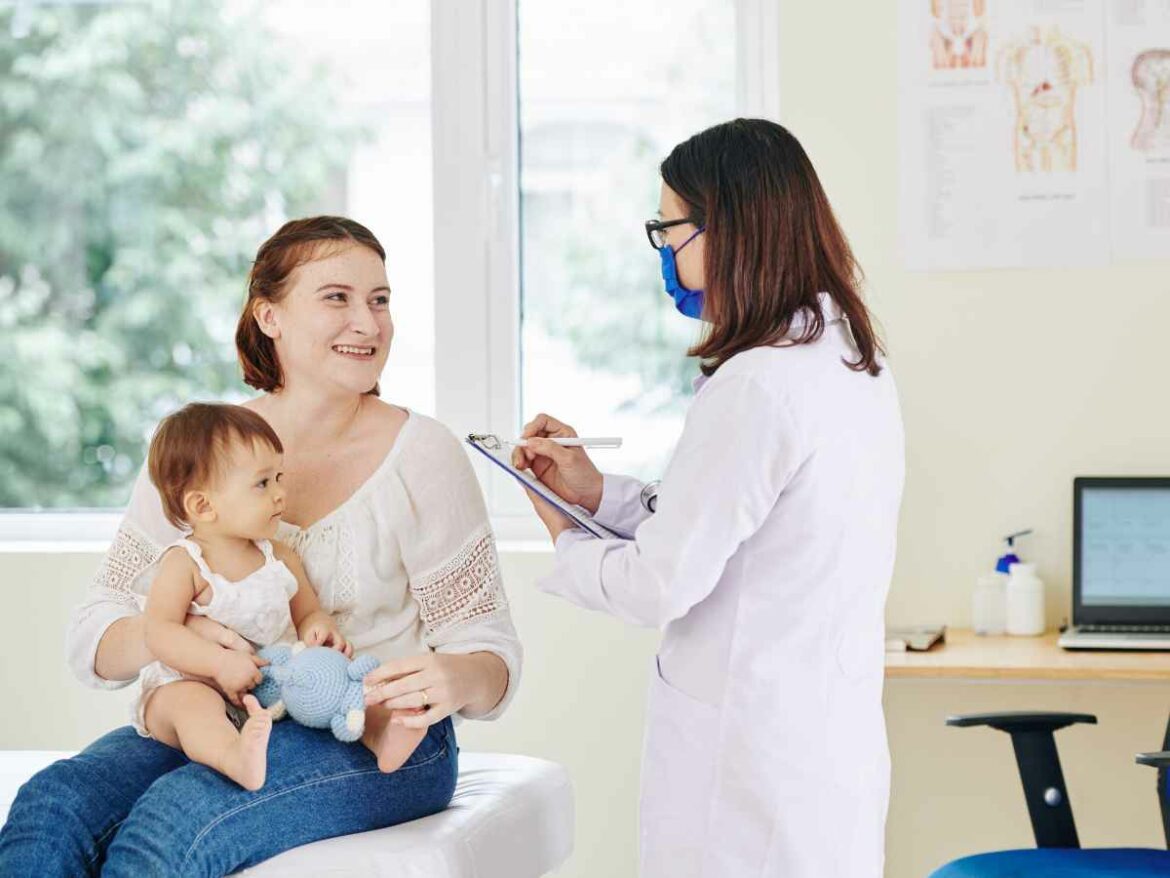 Mãe com bebê no colo conversa com pediatra usando máscara em consultório iluminado. A mãe parece sorridente enquanto tira dúvidas.