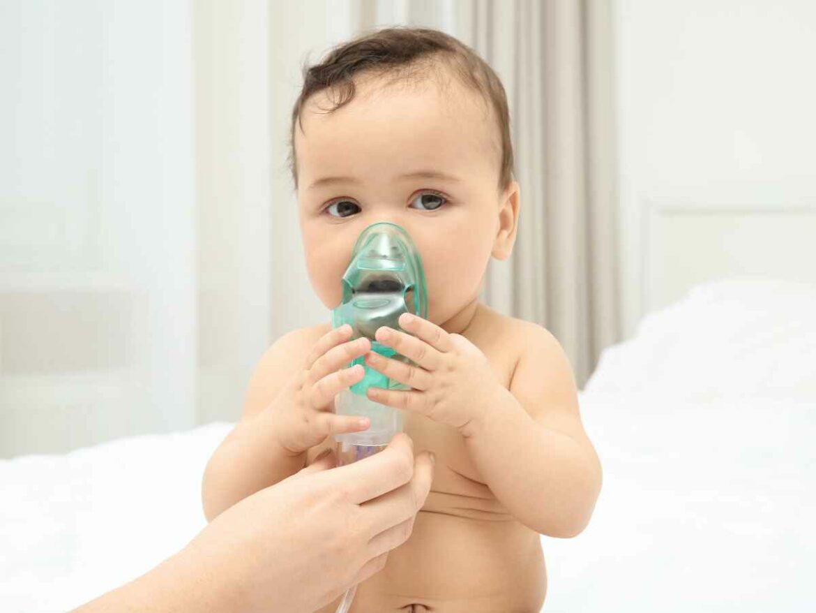 Bebê sentado em ambiente claro segura uma máscara respiratória enquanto recebe ajuda de um adulto durante uma inalação.