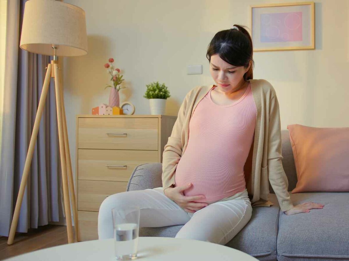 Mulher grávida sentada no sofá com expressão de incômodo, apoiando a mão sobre a barriga, demonstrando desconforto causado por sintomas comuns da gestação.