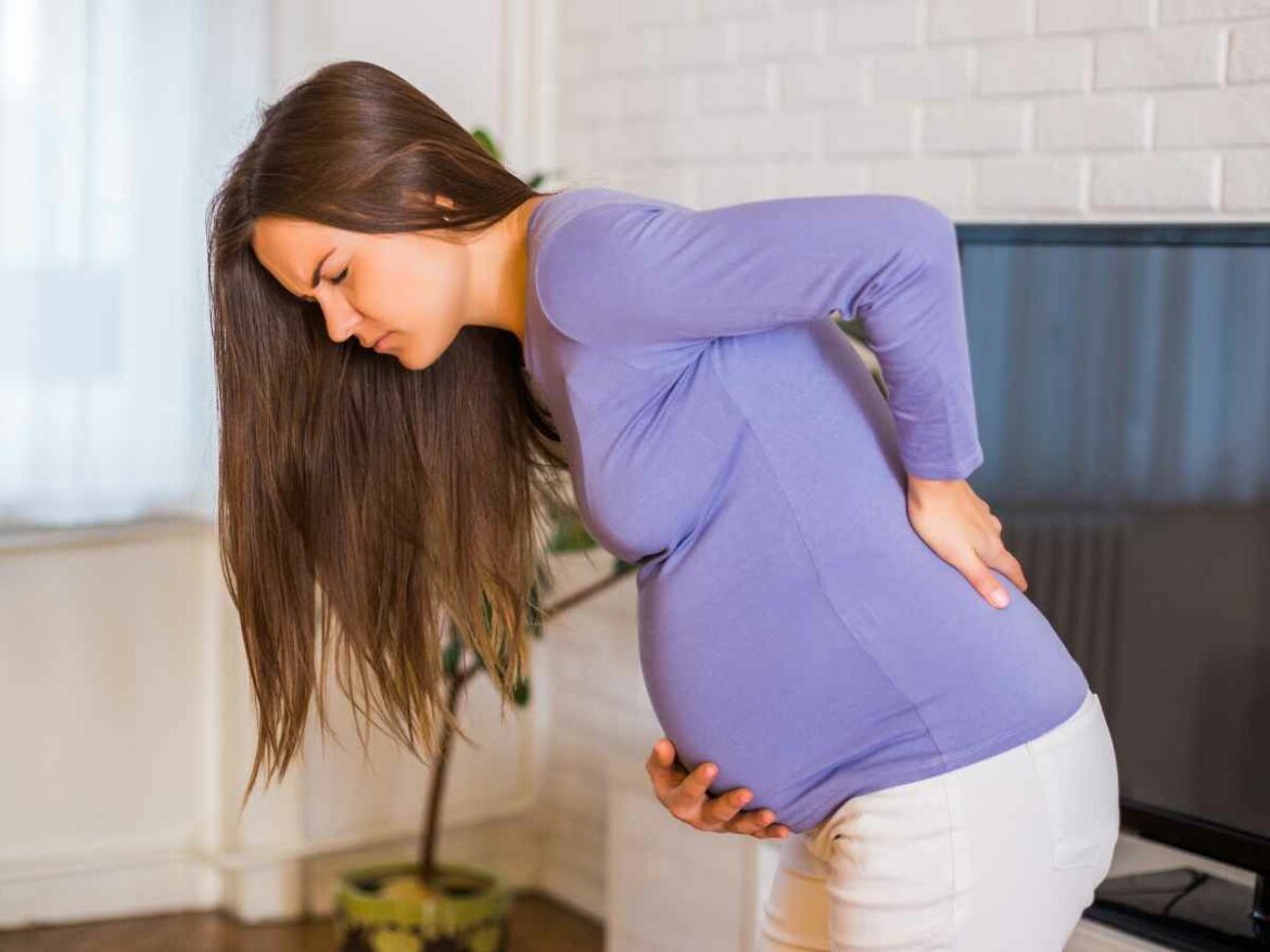 Mulher grávida com expressão de dor enquanto segura a barriga com uma mão e apoia as costas com a outra, inclinando-se levemente para frente, demonstrando desconforto comum.