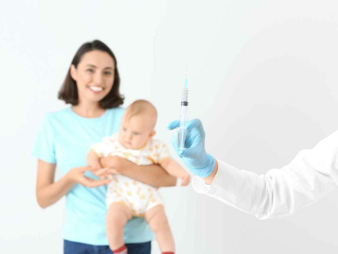Mãe sorri ao fundo segurando o bebê no colo, enquanto profissional da saúde em primeiro plano segura uma seringa com luvas azuis.