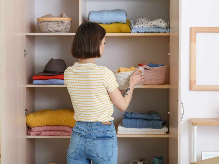 Mulher organiza prateleiras de um armário com roupas de bebê dobradas e cestos organizadores, transmitindo cuidado.
