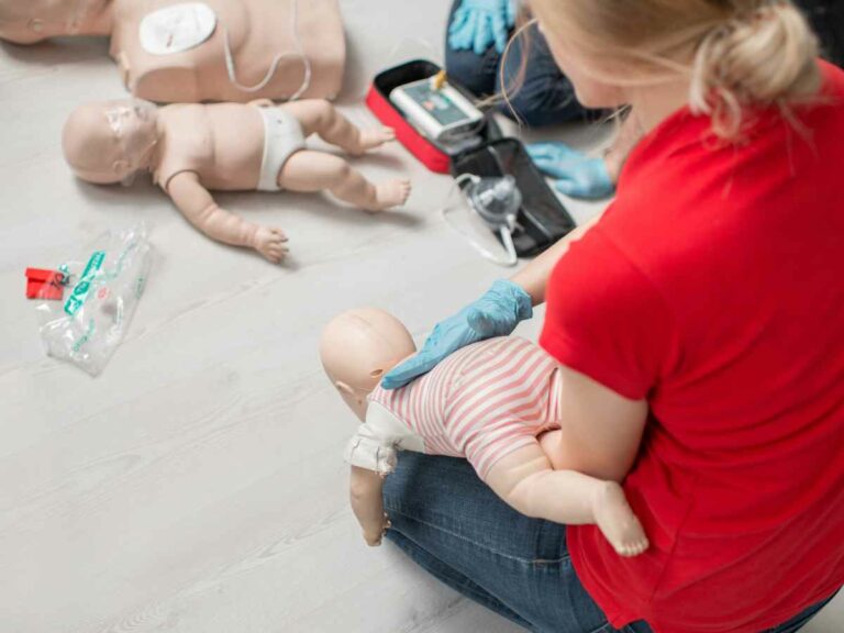 Pessoa realiza treinamento de primeiros socorros em boneco de bebê, simulando manobra de desobstrução das vias aéreas, com outros bonecos e equipamentos médicos ao redor.