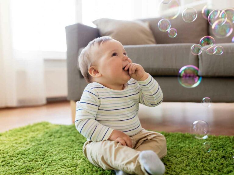 Bebê loiro sentado em um tapete verde dentro de casa, vestindo blusa listrada e calça bege, observando e brincando com bolhas de sabão coloridas no ar.