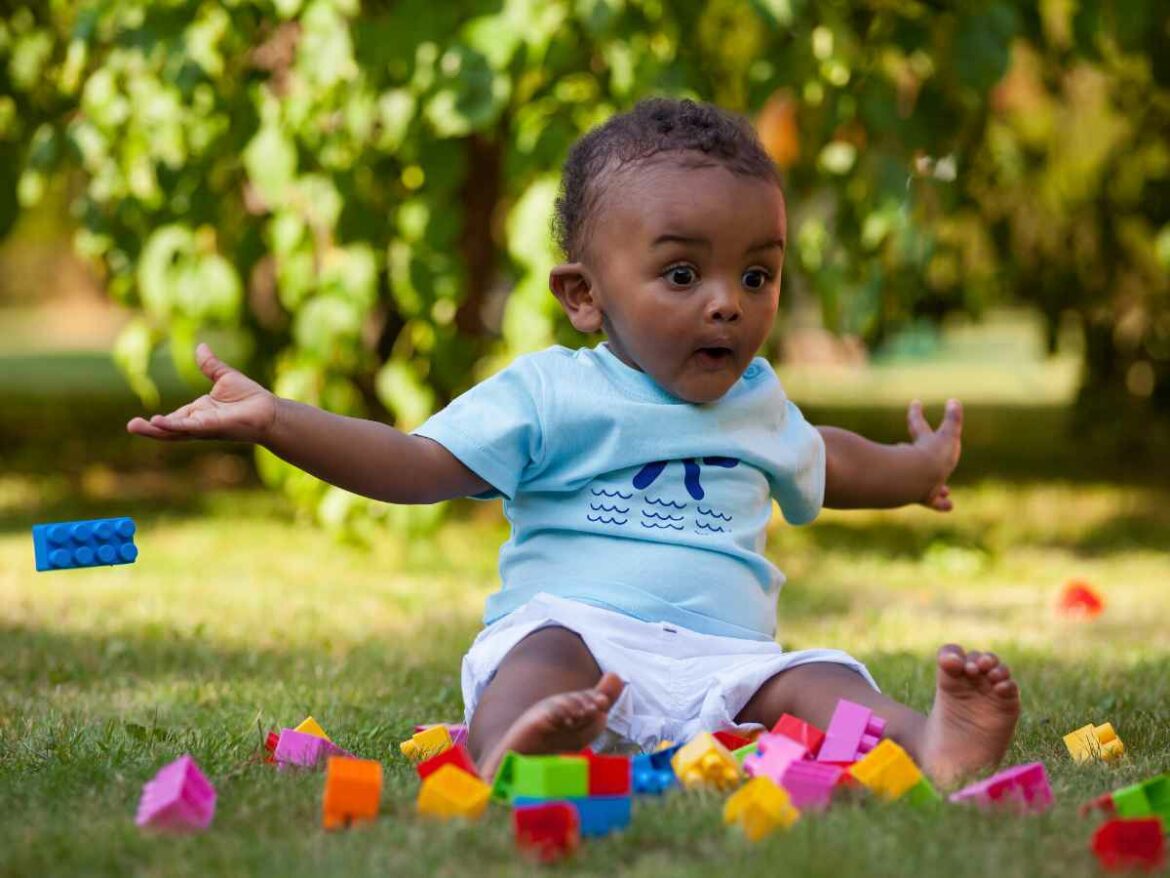 Bebê sentado na grama, usando camiseta azul clara, cercado por blocos de montar coloridos, com expressão de surpresa enquanto brinca.