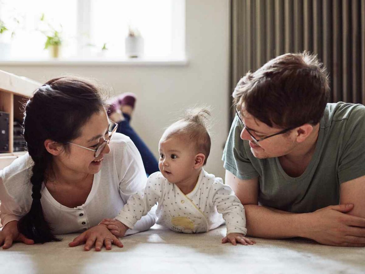 Casal de pais deitados no chão de casa sorrindo para o bebê, que está engatinhando entre eles com expressão curiosa. A cena transmite carinho, cuidado e vínculo familiar.