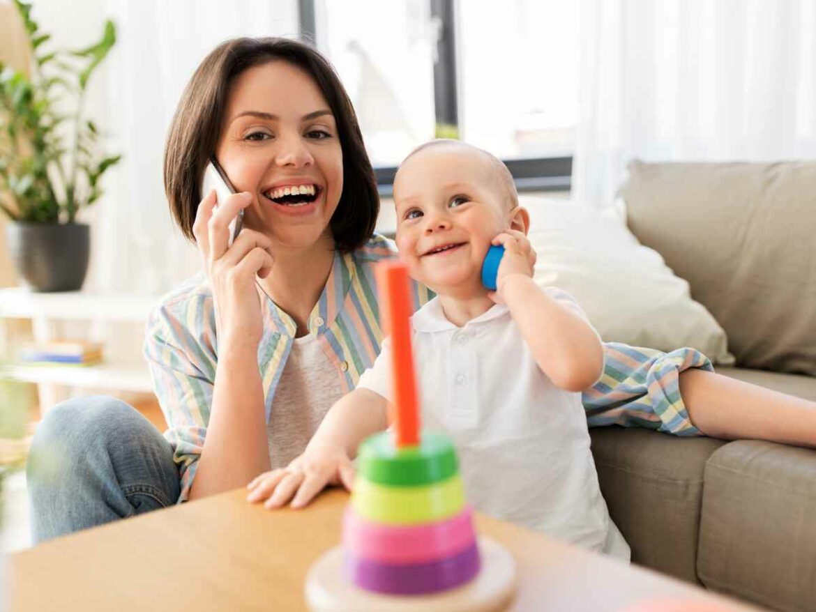 Mãe e bebê se divertem juntos no sofá enquanto brincam de falar ao telefone. A mãe segura um celular de verdade e sorri, enquanto o bebê imita a ação com uma peça colorida de brinquedo junto ao ouvido.