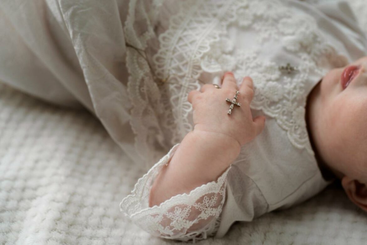 Bebê deitada na cama com roupa de batismo segurando um colar com crucifixo.