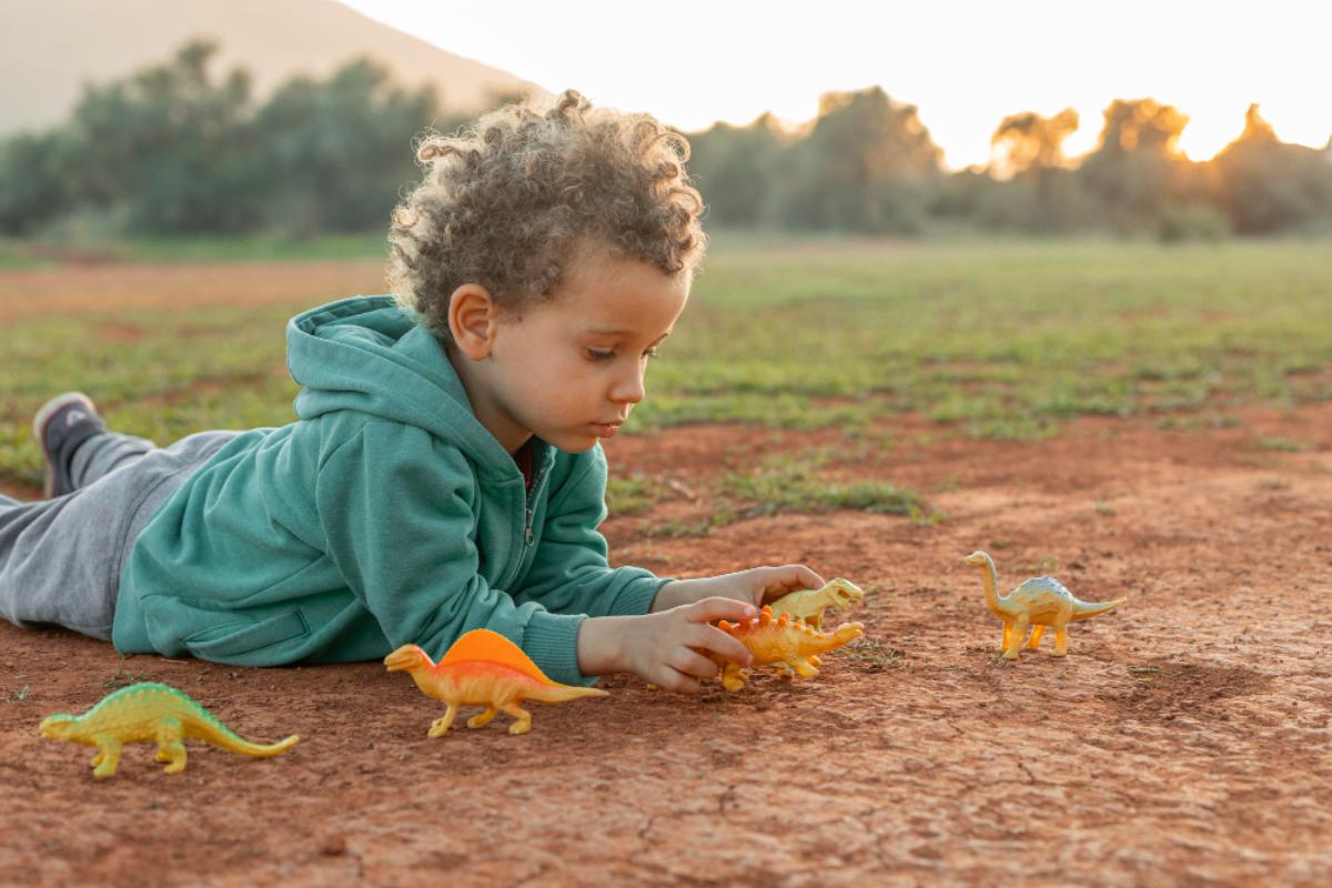 Criança deitada no chão de terra, ao fundo gramado. O menino está com moletom verde e calça cinza e está brincando com três dinossauros de brinquedo.