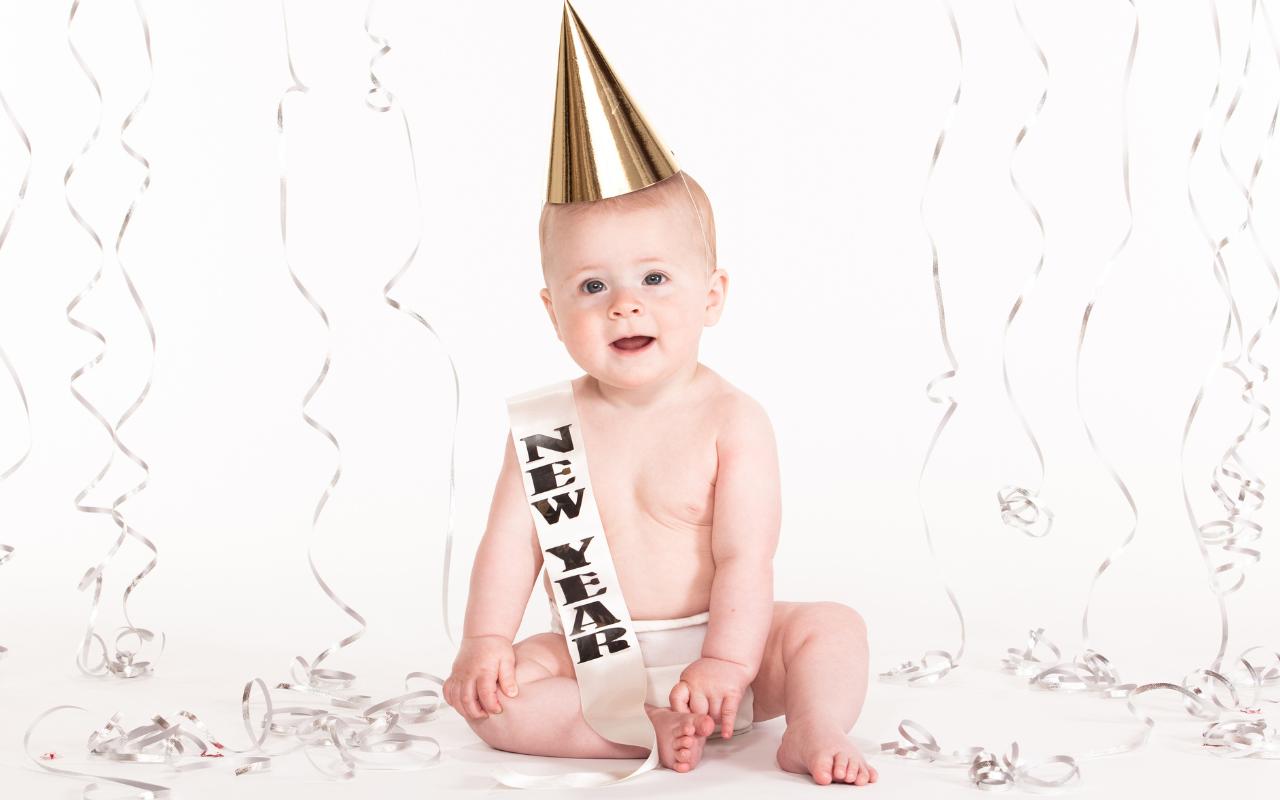 Bebê sentado em fundo branco com confetes e com uma faixa de "new year" e chapéu de festa dourado.