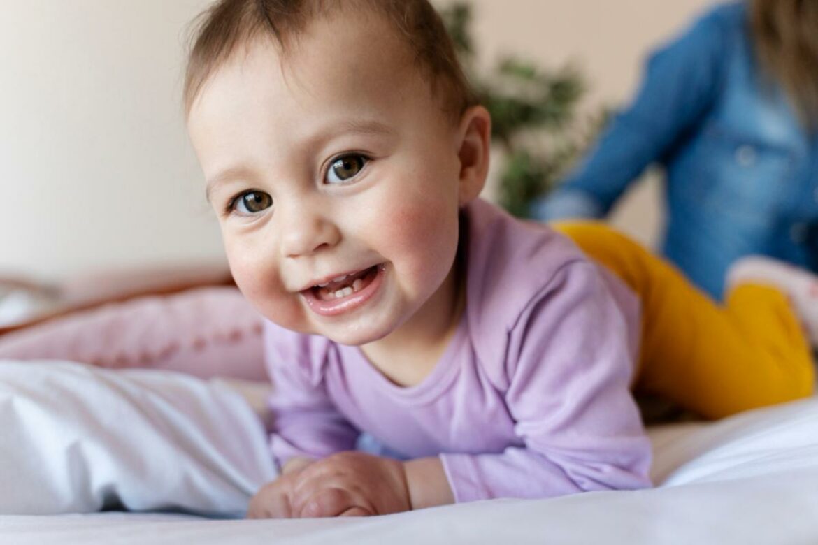 Bebê deitada na cama e sorrindo, mostrando os dentes.