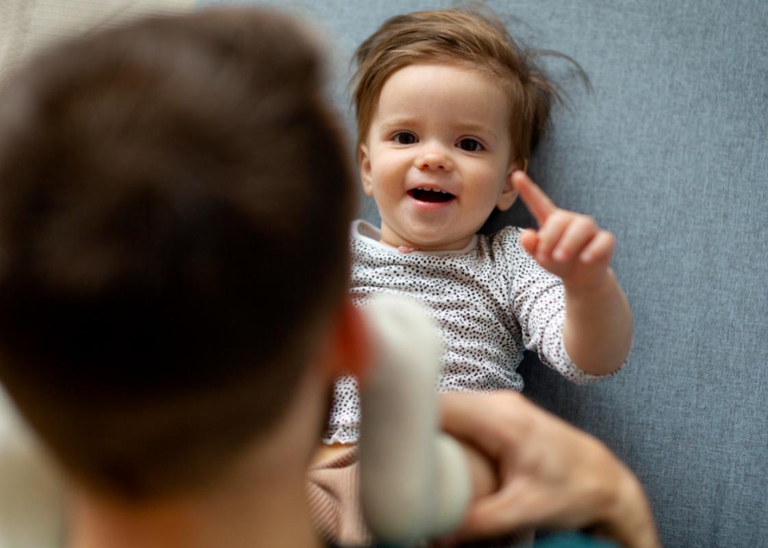 Bebê deitado olhando e apontando para cima enquanto pai observa e brinca.