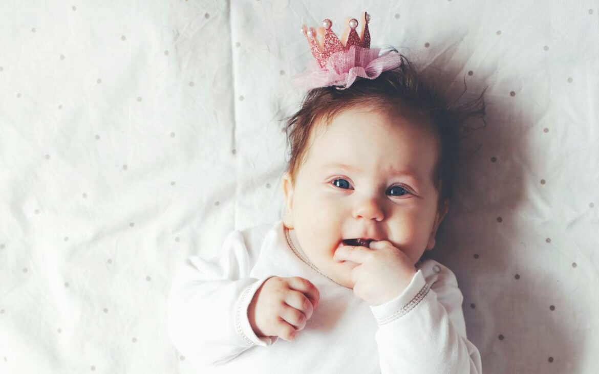 Bebê deitada na cama com coroa de princesa e roupinha branca.