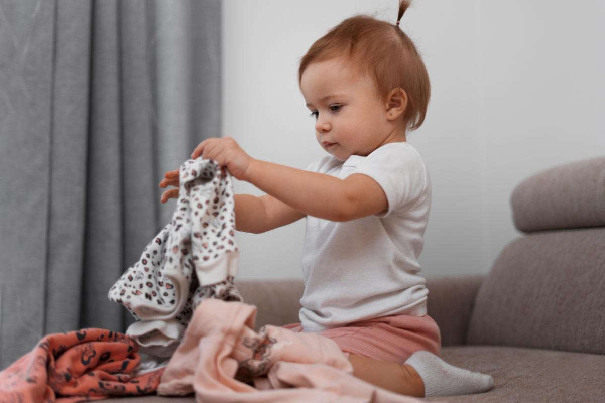 Bebê sentada na cama brincando com as roupas que ganhou de presente no Dia das Crianças.