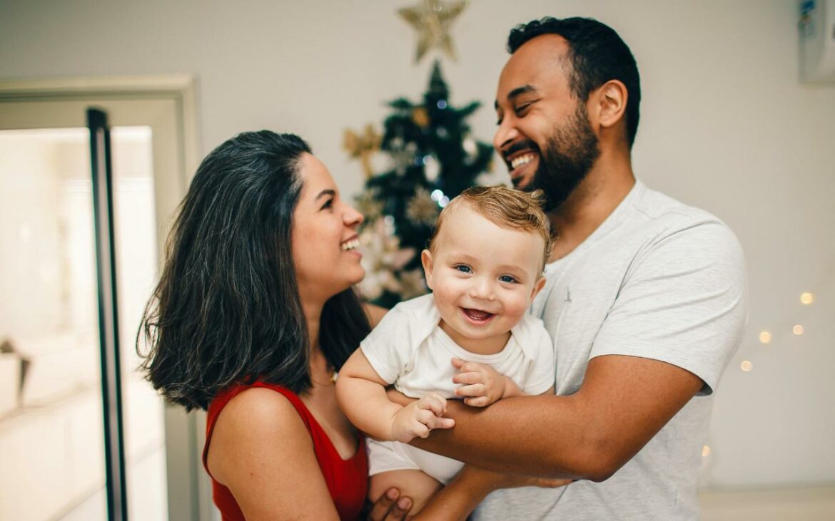 Mãe, pai e bebê sorrindo e brincando em frente à arvore de Natal.