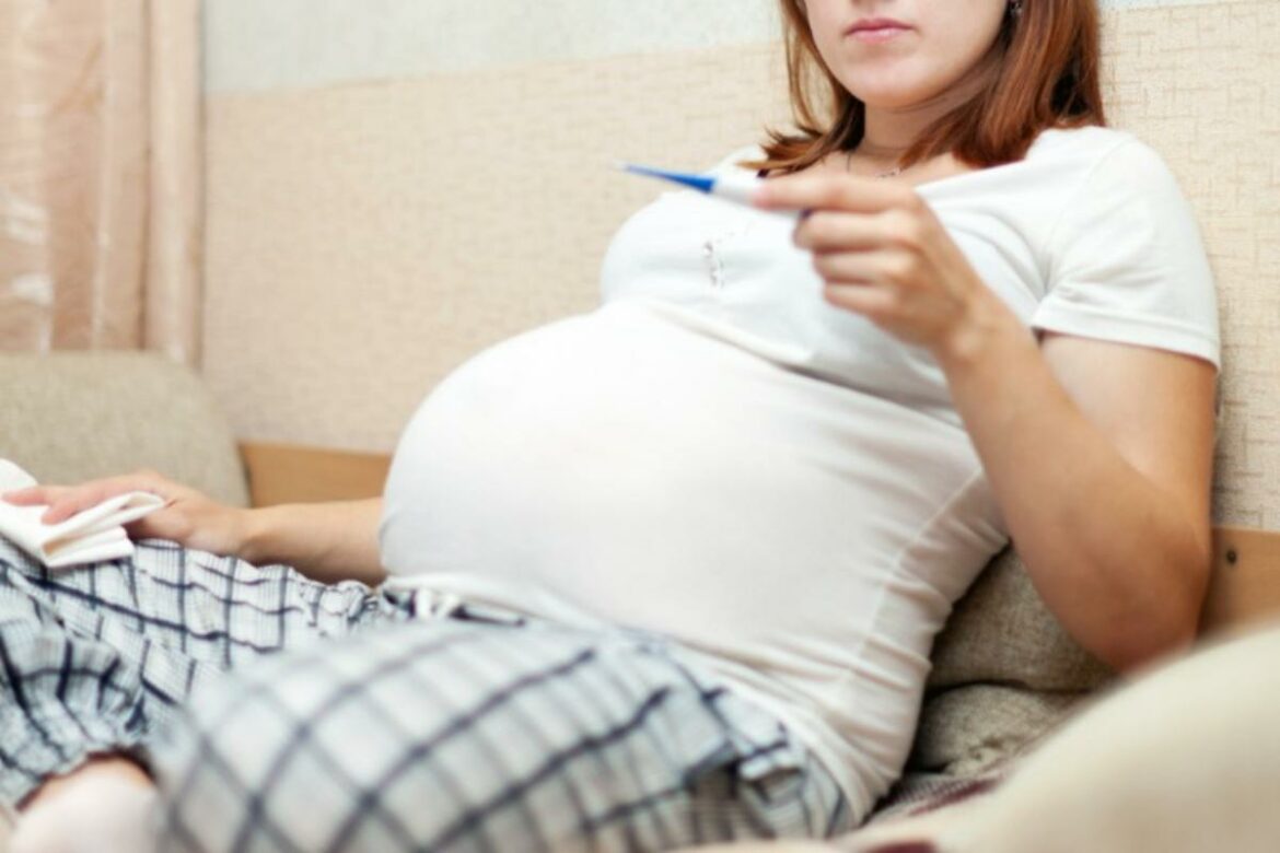 Grávida sentada no sofá conferindo o termômetro.