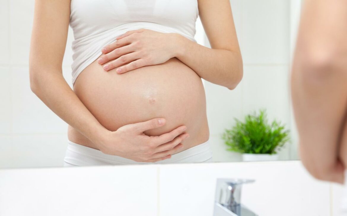 Mulher grávida em frente ao espelho do banheiro segurando a barriga.