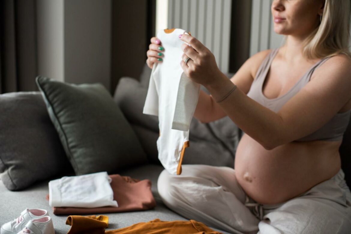 Gestante sentada na cama dobrando uma roupa de bebê. Ao seu lado tem mais uma pequena pilha de roupas infantis.