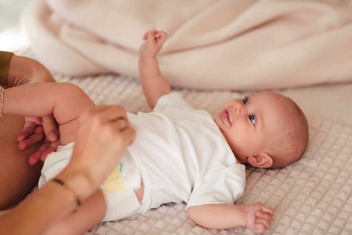Mãe trocando bebê que está deitando na cama sorrindo.