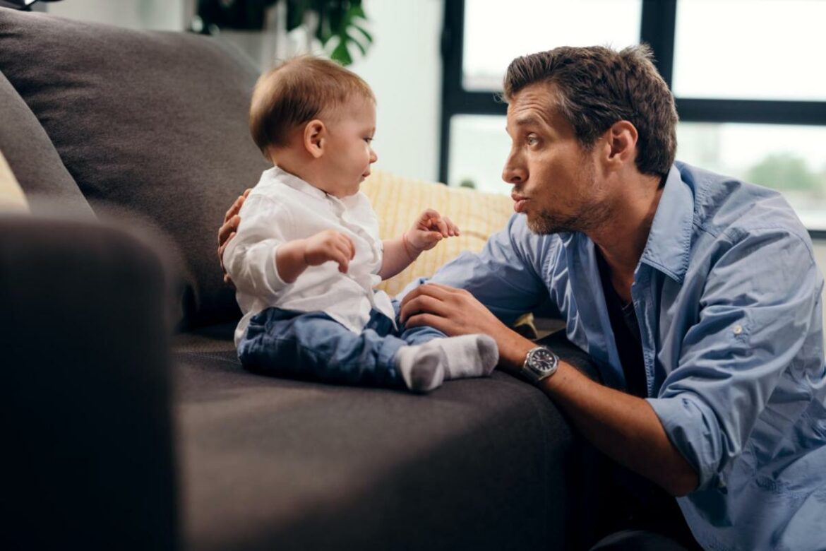 Pai conversando com bebê que está sentado no sofá.