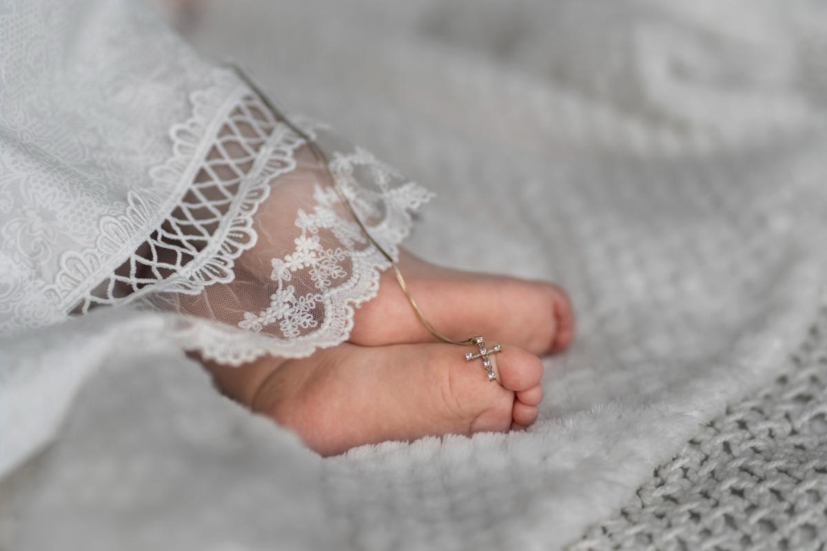 Bebê deitado apenas com os pés e a barra do vestido de batizado aparecendo. Entre os pés, colar dourado com pingente de cruz.