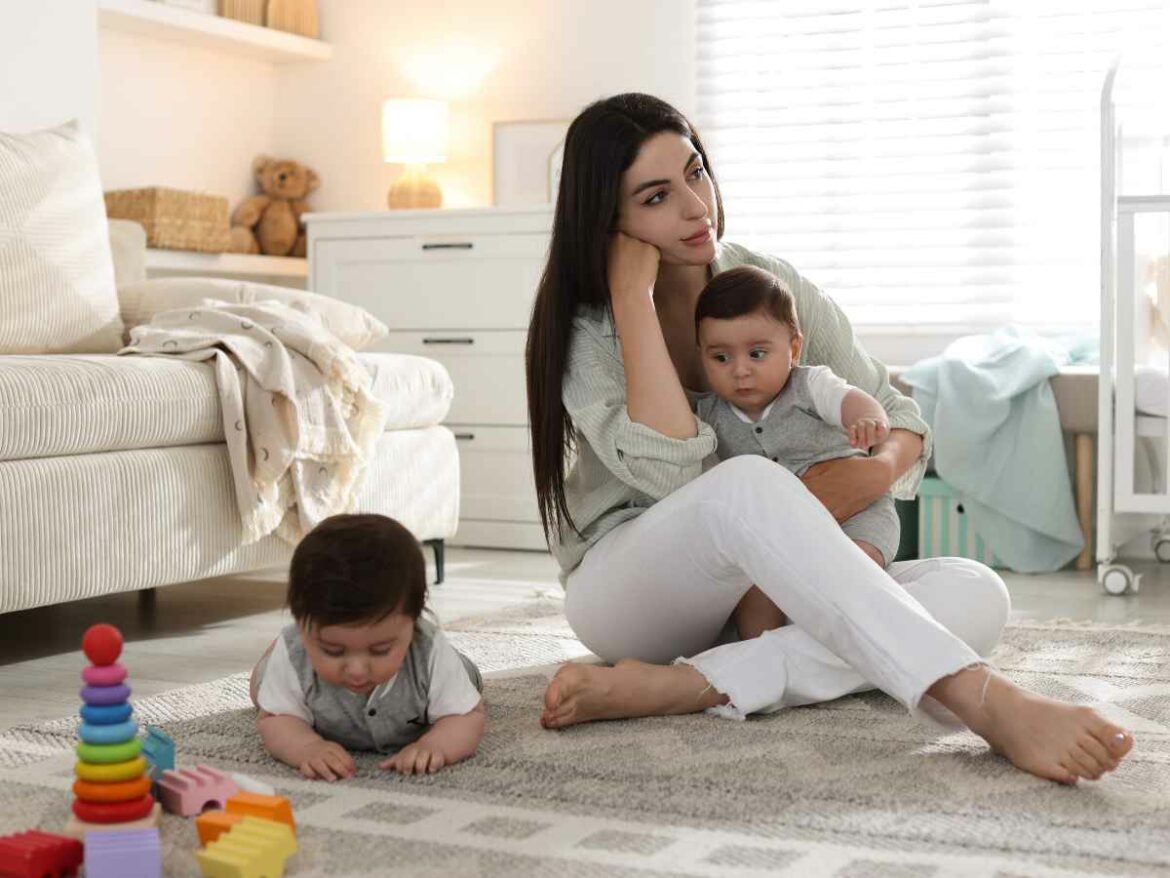 Mãe sentada no chão de um quarto infantil, com expressão de cansaço, segurando um bebê no colo enquanto outro brinca no tapete com brinquedos coloridos