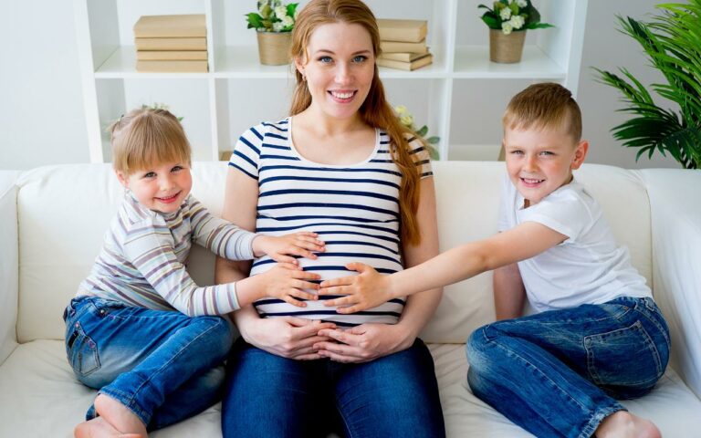 Mulher sentada no sofá grávida pela terceira vez ao lado dos filhos.