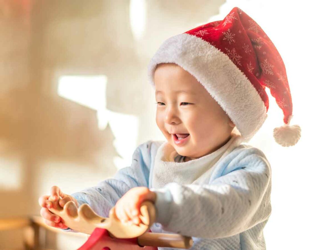 Bebê usando gorro de Natal e sorrindo enquanto segura brinquedo de madeira em ambiente iluminado pela luz natural.