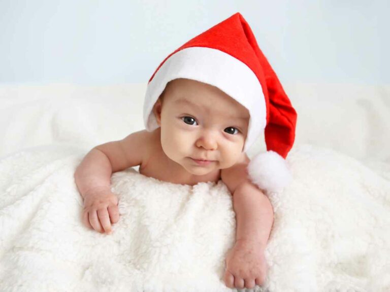 Bebê deitado sobre manta branca usando gorro de Natal vermelho, olhando para a câmera em ambiente claro e aconchegante.