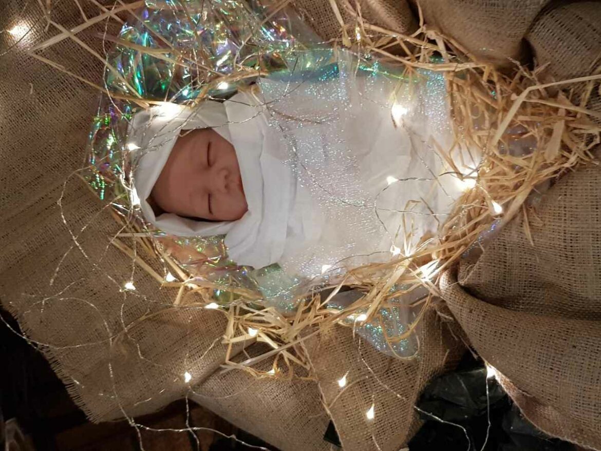 Bebê dormindo envolto em tecido branco, posicionado em cenário decorado com luzes natalinas e elementos que remetem ao presépio.