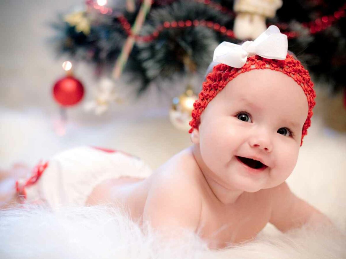 Bebê sorrindo de bruços sobre superfície macia, usando faixa vermelha com laço branco em cenário decorado para o Natal.