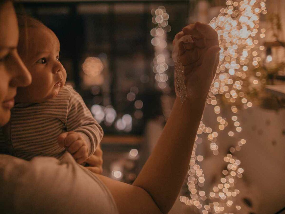 Mãe segurando um bebê no colo enquanto ambos observam luzes de Natal acesas em ambiente interno com iluminação suave.
