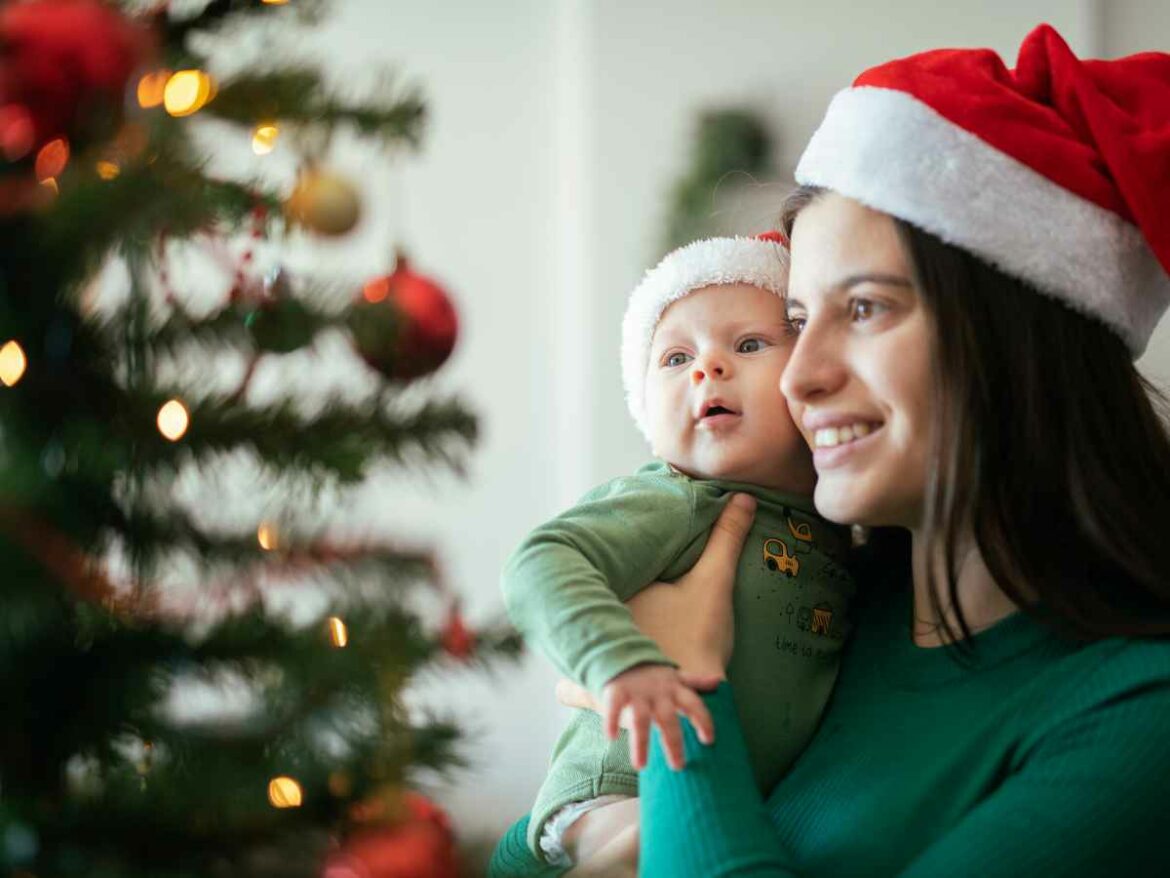 Mulher segurando um bebê no colo próximo a uma árvore de Natal decorada, ambos usando gorros natalinos.