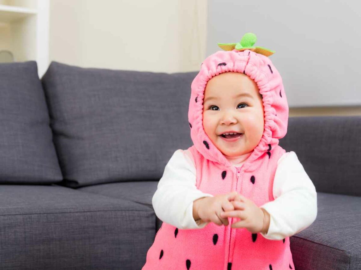 Bebê sorridente usando fantasia rosa de melancia, sentado em um sofá cinza