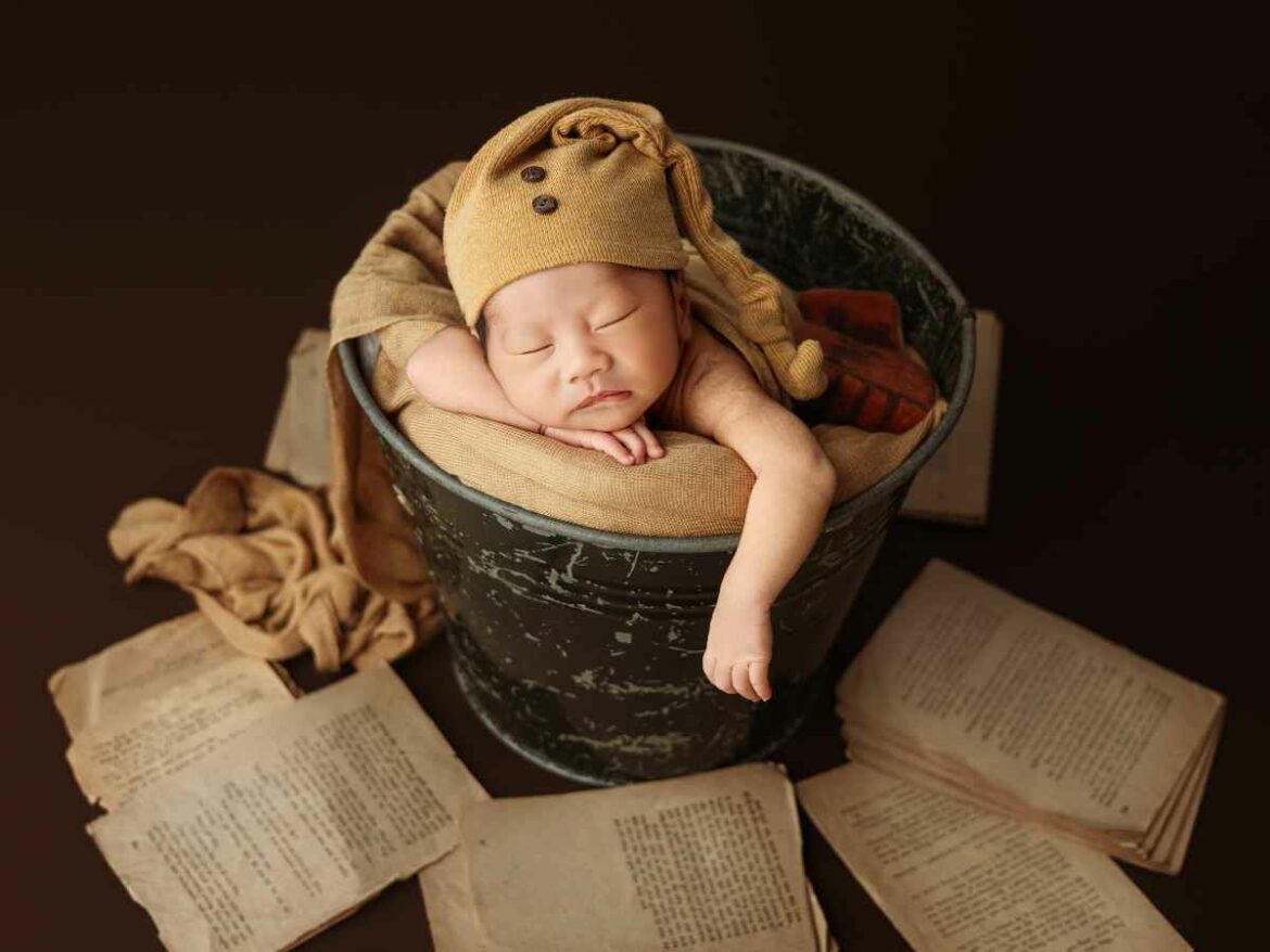 Bebê recém-nascido dormindo dentro de um balde decorativo, envolto em tecido bege, com páginas da biblia ao redor