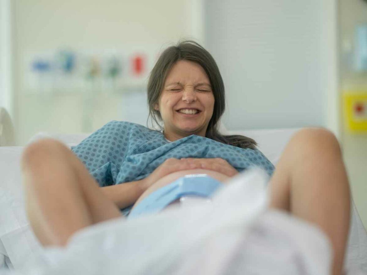 Mulher deitada em maca hospitalar usando camisola médica, com expressão de esforço e dor durante o trabalho de parto.