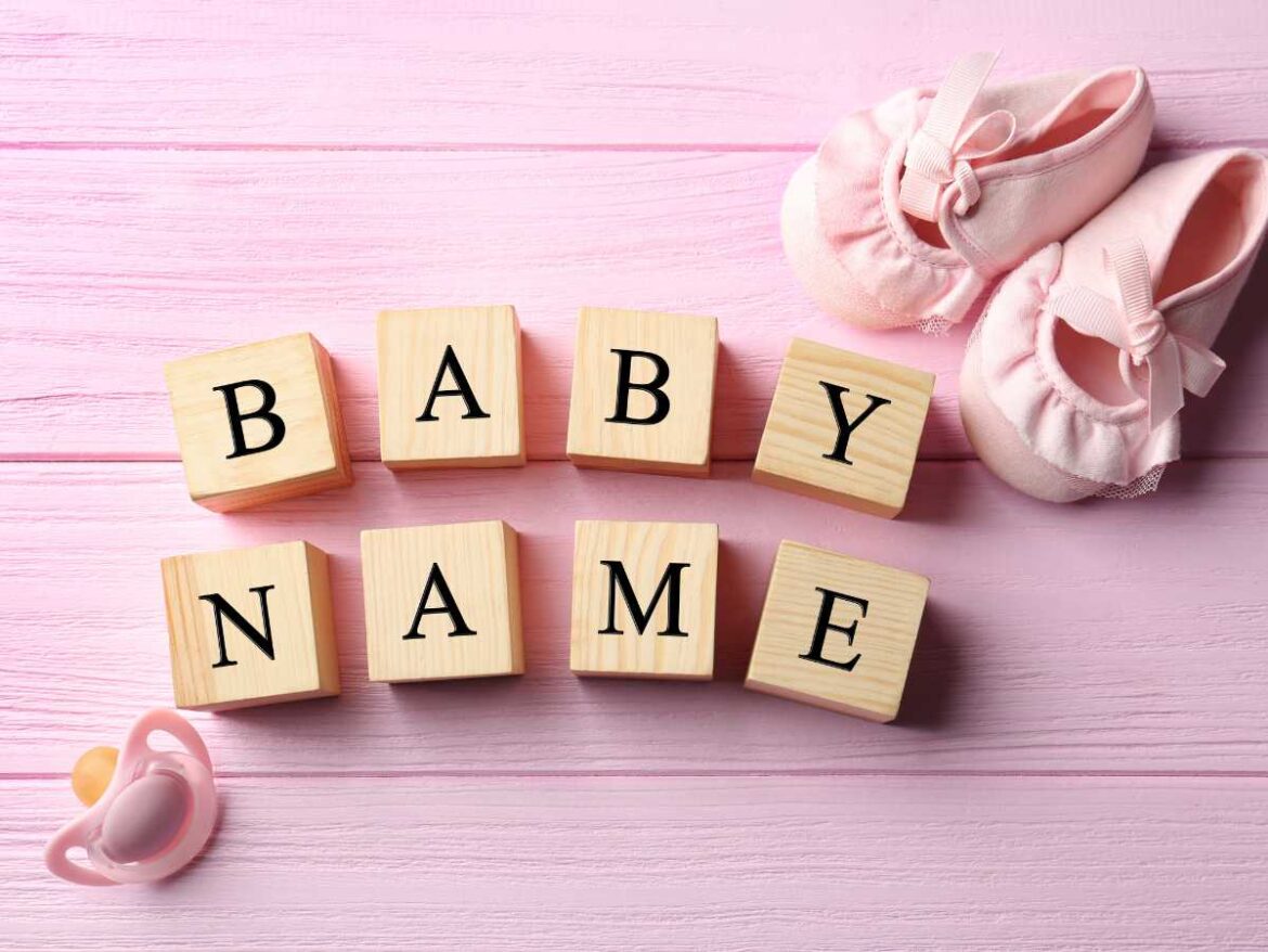 Blocos de madeira formando a palavra “baby name” ao lado de sapatinhos e chupeta sobre fundo rosa