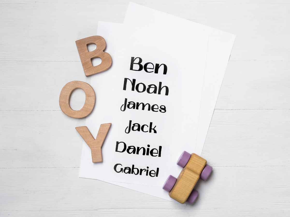 Lista de nomes masculinos como Ben, Noah, James, Jack, Daniel e Gabriel ao lado da palavra “boy” em letras de madeira e um carrinho de brinquedo, representando a escolha do nome do bebê.