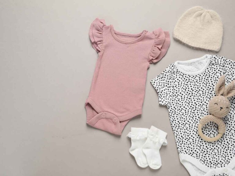 Conjunto de roupas delicadas para bebê, incluindo body rosa, body estampado, meias e touca, ao lado de brinquedo macio