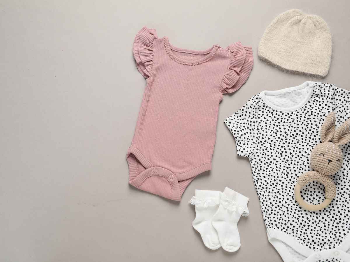Conjunto de roupas delicadas para bebê, incluindo body rosa, body estampado, meias e touca, ao lado de brinquedo macio